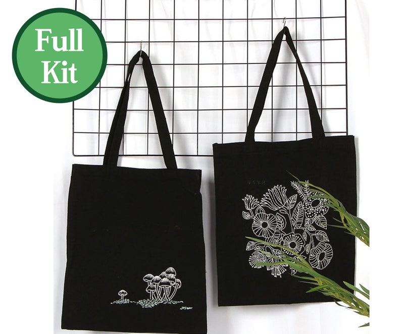 May include: Two black tote bags with white embroidery. One bag features a mushroom design, the other features a floral design. The bags are hanging on a black wire grid.