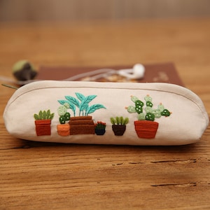May include: A white canvas pencil case with embroidered green plants in brown pots. The plants include a cactus, a succulent, and a leafy plant.