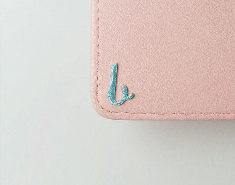 May include: A light pink leather wallet with a light blue embroidered letter "L" on the front.