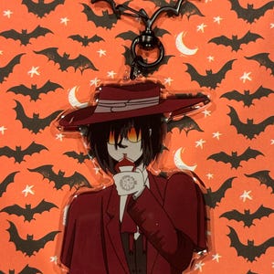 May include: A Halloween-themed keychain featuring a character in a red suit and hat, with a bat-shaped clasp. The character has yellow eyes and is holding a cup. The background is orange with bat and moon patterns.