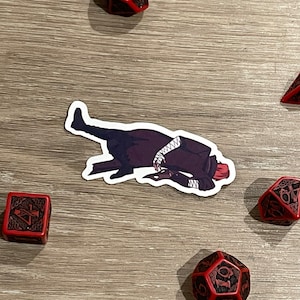 May include: A sticker of a person lying down, dressed in a dark coat and white accents, surrounded by red and black dice with intricate designs. The dice are in various shapes, including a D20.