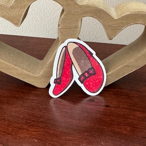 Może przedstawiać: Naklejka przedstawiająca parę czerwonych butów z kokardkami. Buty mają brązowe wnętrze i są umieszczone na drewnianym tle w kształcie serca. Buty są w żywym czerwonym kolorze.