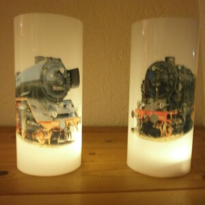May include: Two cylindrical, white candle holders featuring images of vintage steam trains. The trains are detailed with black, blue, orange, and silver accents. The candle holders are lit from within, producing a warm glow.