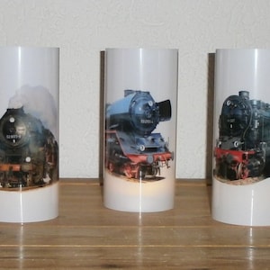 May include: Three white cylindrical candle holders with images of vintage steam locomotives. The locomotives are black, blue, and red, with smoke billowing from the stacks. The candle holders are arranged on a wooden surface.
