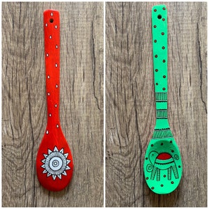 Könnte beinhalten: Zwei bemalte Holzlöffel mit unterschiedlichen Designs. Der erste Löffel ist rot mit weißen Punkten und einem weißen Mandala-Design. Der zweite Löffel ist grün mit roten Punkten und einem schwarz-weißen Elefanten-Design.