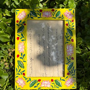 May include: A rectangular yellow picture frame featuring a hand-painted border of pink lotus flowers, green leaves, and small red buds. The frame contains a piece of paper with handwritten text in a script that appears to be Devanagari.