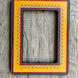 Op de afbeelding: Een zwart houten fotolijst met een gele en rode rand. De lijst heeft een geometrisch patroon rond de rand.