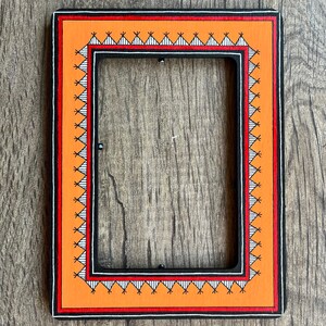 Op de afbeelding: Een zwart fotolijstje met een oranje rand en ingewikkelde geometrische ontwerpen in wit en zwart. Het lijstje is gemaakt van hout en heeft een rechthoekige vorm.