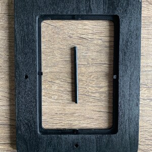 Op de afbeelding: Zwarte houten lijst met een rechthoekige uitsparing. Een kleine zwarte metalen staaf bevindt zich in het midden van de lijst.