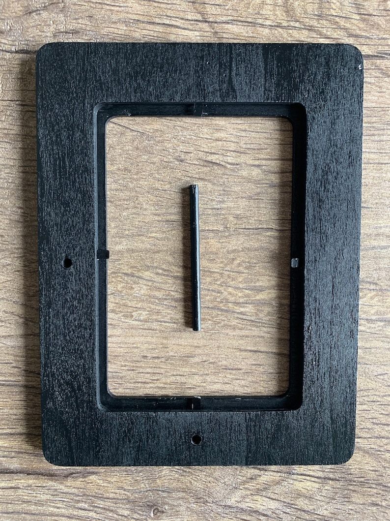 Op de afbeelding: Zwarte houten lijst met een rechthoekige uitsparing. Een kleine zwarte metalen staaf bevindt zich in het midden van de lijst.