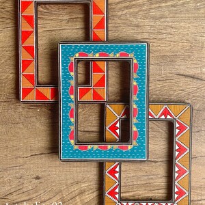 May include: Three wooden picture frames with intricate hand-painted designs. The frames are nested inside each other, with the largest frame featuring a red, orange, and yellow geometric pattern, the middle frame has a blue and white pattern, and the smallest frame has a red, orange, and yellow pattern.