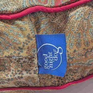 May include: Close-up of a dog bed with a paisley pattern in shades of gold, teal, and red. A blue tag with the words "good night dog" is attached to the bed. The bed has a red trim.