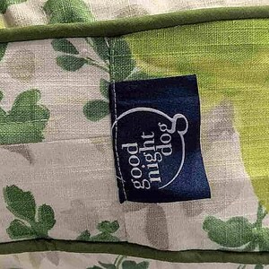 May include: Close-up of a dog bed with a floral pattern in shades of green and white. A dark blue tag with the words "good night dog" in white is attached to the bed.