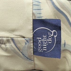 May include: Close-up of a dog bed with a blue label that says "good night dog" in white script. The bed has a cream-colored fabric with a blue and gray pattern. The label is sewn onto the bed.