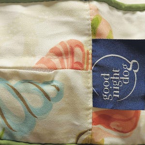 May include: Close-up of a dog bed with a floral pattern in shades of blue, peach, and green. A navy blue tag with the words "good night dog" in white is sewn onto the bed.