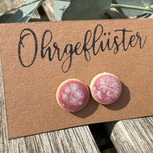 Könnte beinhalten: Runde Ohrringe mit rosa und weiß gesprenkeltem Muster, präsentiert auf einer braunen Karte. Auf der Karte steht das Wort "Ohrgeflüster" in schwarzer Schrift. Die Ohrringe liegen auf einer Holzoberfläche mit etwas Grün im Hintergrund.