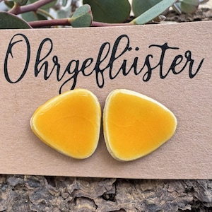 Könnte beinhalten: Ein Paar gelbe, dreieckige Ohrringe auf einer braunen Karte mit dem Wort "Ohrgeflüster" in schwarzer Schrift. Die Ohrringe sind leuchtend gelb und haben eine glatte, polierte Oberfläche. Die Karte ist vor einem natürlichen Holzuntergrund platziert.