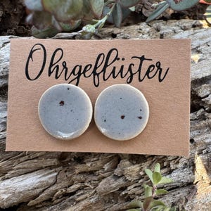 Könnte beinhalten: Ein Paar hellblaue Keramik-Ohrstecker mit dunklen Sprenkeln, präsentiert auf einer braunen Karte mit dem Wort "Ohrgeflüster" in schwarzer Schrift. Die Ohrringe sind rund und liegen auf einem Stück verwittertem Holz.
