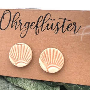 Könnte beinhalten: Ein Paar cremefarbene Keramikohrringe mit einem erhabenen Sonnenstrahlen-Design in hellorange. Die Ohrringe werden auf einer braunen Karte mit dem Text "Ohrgeflüster" in Schwarz präsentiert.