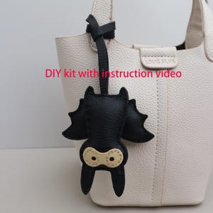 May include: A black leather bat-shaped keychain with beige eye details hangs from a white leather handbag. The keychain is attached to a black leather strap. Text on the image reads: DIY kit with instruction video.
