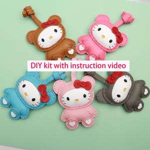 May include: Five colorful leatherette keychains in the shape of a cartoon cat with a red bow. The keychains are brown, pink, blue, light pink, and gray. The text "DIY kit with instruction video" is displayed on the image.
