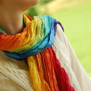 Könnte beinhalten: Ein Regenbogen-Schal, der um den Hals drapiert ist. Die Farben des Schals reichen von Rot bis Violett. Der Schal besteht aus einem weichen, fließenden Stoff. Die Person trägt ein cremefarbenes Hemd.