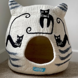 May include: A cream-coloured felted pet cave shaped like a cat head with two ears. It features three black cat silhouettes with white dotted details and long, curved tails. Dark grey stripes accent the sides. A blue label reads EVEREST PET CAVE.