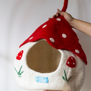 May include: A whimsical pet cave shaped like a mushroom. The cave features a red top with white polka dots and a white base adorned with embroidered mushroom details. The label reads "Everest Pet Cave".