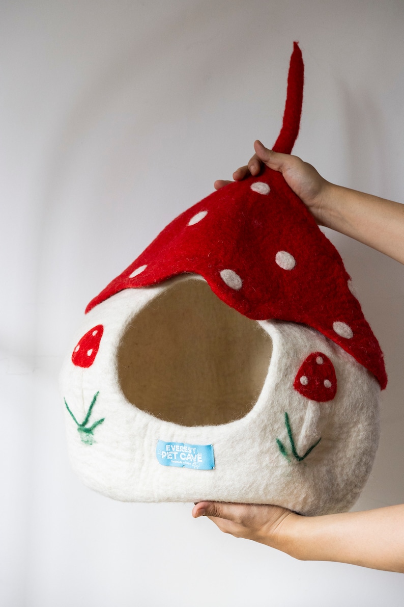 May include: A whimsical pet cave shaped like a mushroom. The cave features a red top with white polka dots and a white base adorned with embroidered mushroom details. The label reads "Everest Pet Cave".