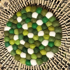 May include: A circular trivet made of felt balls in various shades of green and white. The trivet sits on a woven basket. The felt balls are densely packed together.