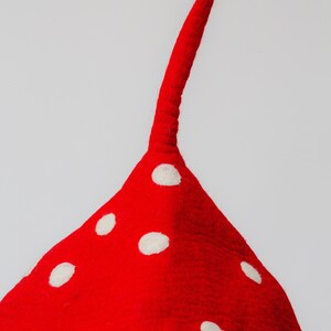 May include: A red felt mushroom-shaped hat with white polka dots. The hat has a long, curved stem extending upwards. The hat is likely handmade and is against a white background.