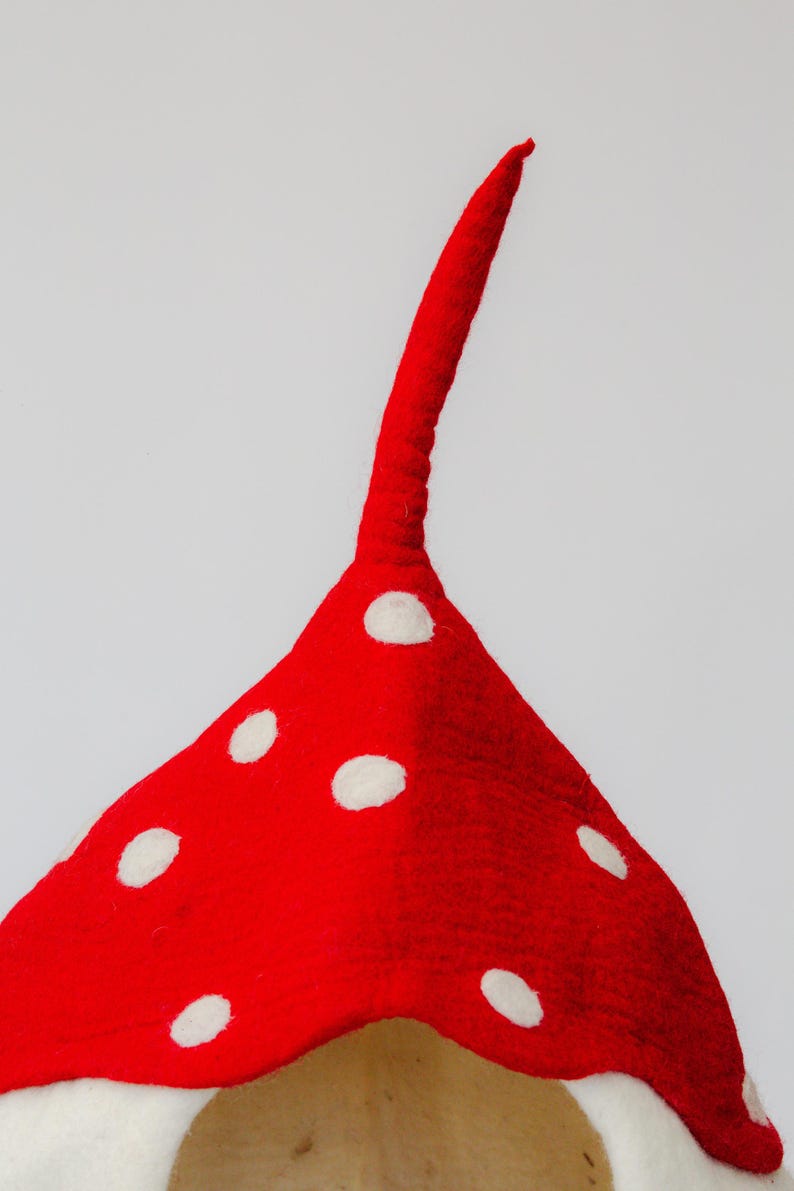 May include: A red felt mushroom-shaped hat with white polka dots. The hat has a long, curved stem extending upwards. The hat is likely handmade and is against a white background.