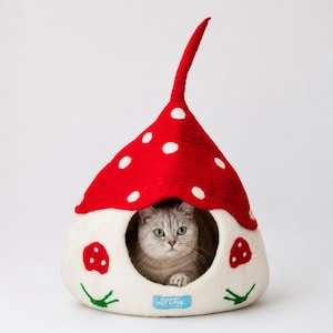 A grey cat inside a Gnome shaped cat cave bed. The bed is white with a red top featuring white spots. The bed has mushroom embroidery on the sides and has a round opening and a small blue tag with text.