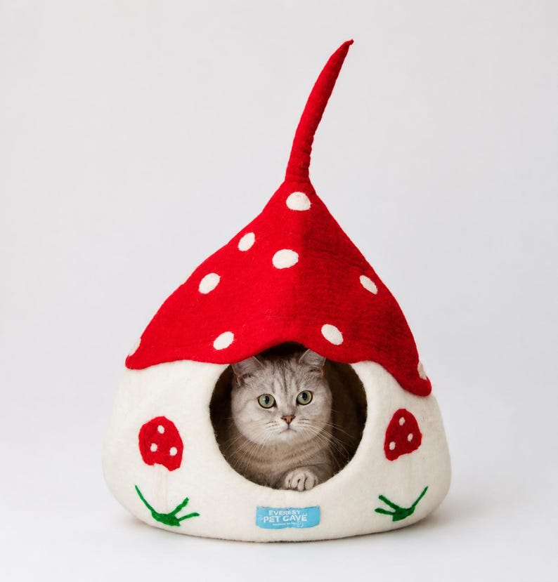 A grey cat inside a Gnome shaped cat cave bed. The bed is white with a red top featuring white spots. The bed has mushroom embroidery on the sides and has a round opening and a small blue tag with text.