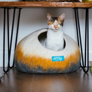 May include: A white and orange cat with closed eyes sits in a gray and orange felt cat bed. The bed has a blue label that reads "Everest Pet Cave".