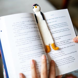 Op de afbeelding: Een handgemaakt pinguïn bladwijzer van wit vilt met zwarte accenten, een roze hoed en oranje voeten. De bladwijzer zit in een open boek. Een uniek en charmant accessoire voor boekenlezers.