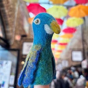 May include: A handmade felt parrot puppet in shades of blue, green, and yellow, with a yellow beak and white accents. The puppet is designed to fit on a finger. The background features colorful umbrellas.