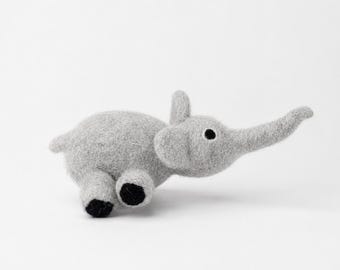 May include: A gray felt elephant toy with black accents. The elephant has a trunk, ears, and small black eyes. The toy is lying on its side, with its legs and tail visible. The elephant is made of a soft, textured material.