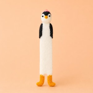 May include: A handmade felt penguin bookmark. The penguin has a white body, black wings, and a pink hat. It has orange feet and a black and white face. The bookmark is designed to be inserted into a book.