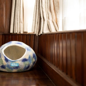May include: A blue and white patterned felt cat bed with a round opening. The bed is sitting on a wooden floor in front of a window.