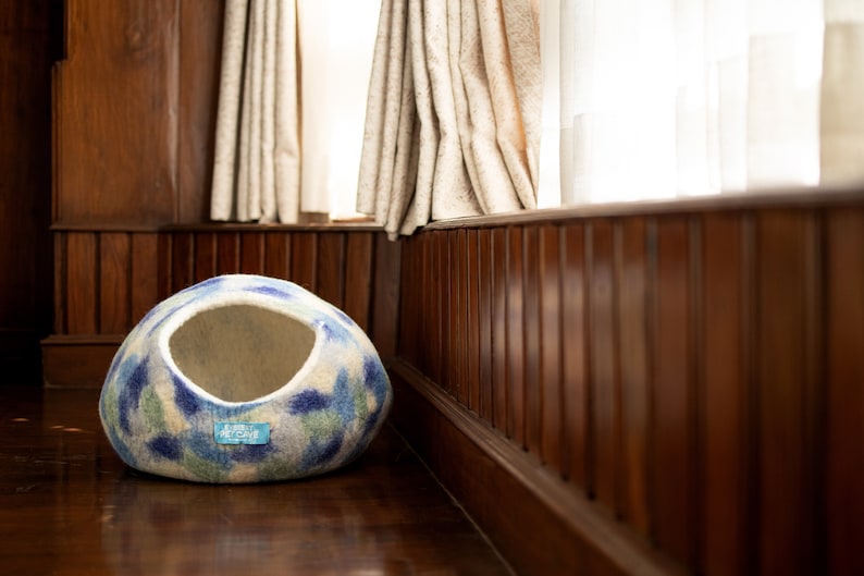 May include: A blue and white patterned felt cat bed with a round opening. The bed is sitting on a wooden floor in front of a window.