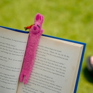 Peut inclure: Marque-page en forme de lapin rose en feutre avec une carotte. Le lapin est placé dans un livre ouvert. Le livre a des bords bleus et est ouvert sur une page de texte.