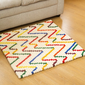 May include: A rectangular, off-white rug with a colorful zigzag pattern. The zigzags are made of small, round, colorful beads in shades of red, orange, yellow, green, blue, and brown. The rug is placed on a wooden floor near a gray sofa.