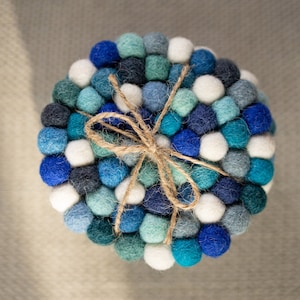 May include: A round felt coaster made of blue, teal, and white pom-poms. The coaster is tied together with a natural twine bow.