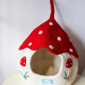 May include: A whimsical pet cave shaped like a mushroom. The red felt roof has white polka dots. The white felt body features a round opening and mushroom accents. The cave has a long, red stem and a blue tag that reads "EVEREST PET CAVE".