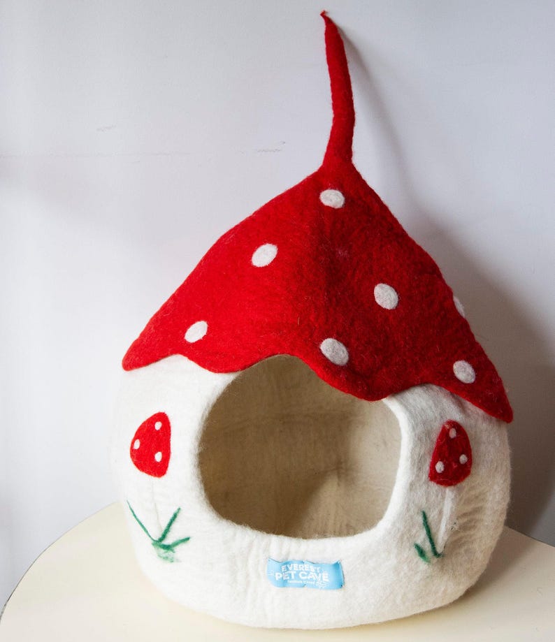May include: A whimsical pet cave shaped like a mushroom. The red felt roof has white polka dots. The white felt body features a round opening and mushroom accents. The cave has a long, red stem and a blue tag that reads "EVEREST PET CAVE".