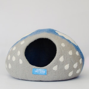 May include: A gray pet cave with a blue top and white raindrop accents. The cave has a large circular opening and a blue label that reads "Everest Pet Cave". The cave is made of felt.