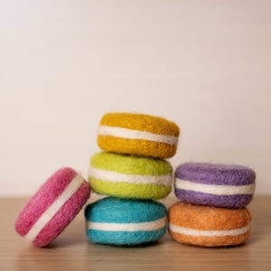 May include: A stack of colorful felt macarons in shades of yellow, green, purple, orange, blue, and pink. Each macaron has a white stripe in the center. The macarons are arranged on a wooden surface.