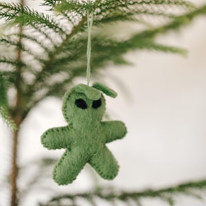 May include: A green felt ornament shaped like a cartoon alien with black eyes hanging from a pine tree branch.