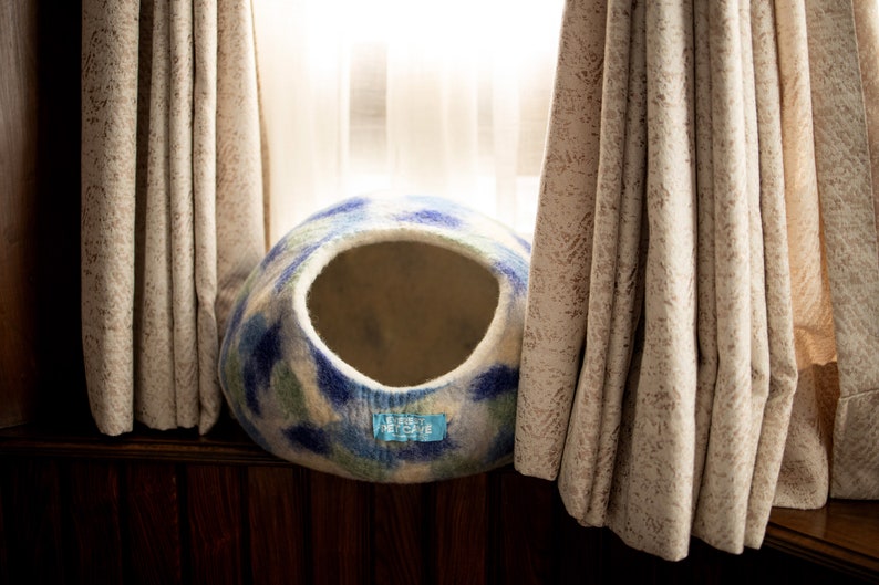 May include: A white, blue and green felted cat bed with a round opening. The bed is sitting on a wooden surface in front of a window with curtains.